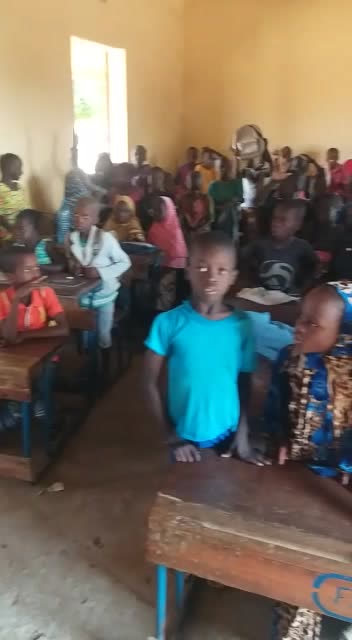 Rows of children at wooden desks fill a classroom, some waving