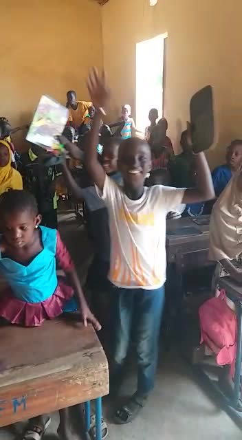 A smiling boy raises a new notebook above his head in a crowded classroom